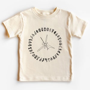 K&ouml;nnte beinhalten: Ein cremefarbenes T-Shirt mit kurzen &Auml;rmeln und einem schwarzen Aufdruck eines Kreises mit Symbolen am Rand und einem Stern in der Mitte.