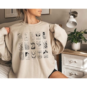 May include: A beige sweatshirt with a black and white graphic print of tarot cards. The cards are arranged in a grid pattern and each card features a different image and title. The titles include "The Eye", "The Ruined", "The Game", "The Celebration", "The End", "The Fool", "The Hive", "The Flame", "The Lovely", "The Slaughterhouse", "The Spiral", "The Stranger", "The Void", "The Wild", "The Extinction".