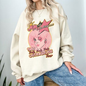 May include: A cream coloured sweatshirt with a vintage-style graphic of Jem and the Holograms. The graphic features a pink and yellow star, a pink circle with a cartoon image of Jem, and the text "Jem and the Holograms", "1987", "Truly, Truly, Truly OUTRAGEOUS Tour".