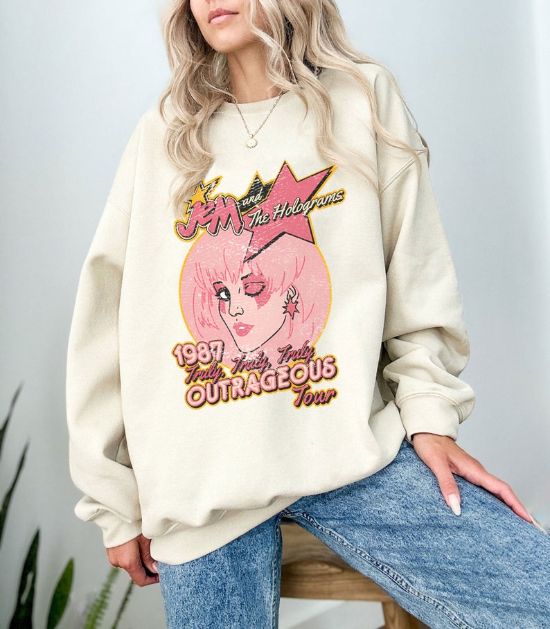 May include: A cream coloured sweatshirt with a vintage-style graphic of Jem and the Holograms. The graphic features a pink and yellow star, a pink circle with a cartoon image of Jem, and the text "Jem and the Holograms", "1987", "Truly, Truly, Truly OUTRAGEOUS Tour".