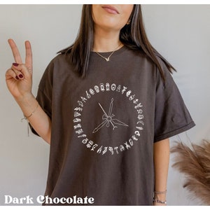 K&ouml;nnte beinhalten: Ein dunkelbraunes T-Shirt mit einem wei&szlig;en Grafikdesign eines Kreises mit einem Kompass und verschiedenen Symbolen am Rand.