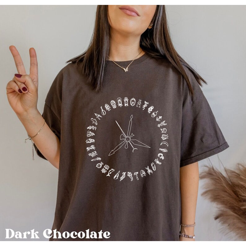 K&ouml;nnte beinhalten: Ein dunkelbraunes T-Shirt mit einem wei&szlig;en Grafikdesign eines Kreises mit einem Kompass und verschiedenen Symbolen am Rand.