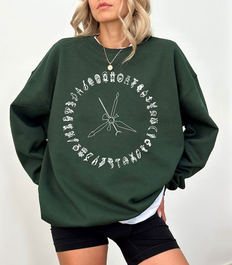 K&ouml;nnte beinhalten: Ein dunkelgr&uuml;ner Pullover mit einem wei&szlig;en Grafikdruck eines Kreises mit verschiedenen Symbolen und einem Paar gekreuzter Schwerter in der Mitte.