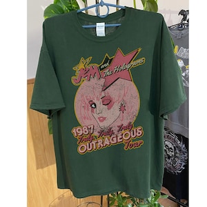 May include: A green t-shirt featuring a vintage-style graphic of Jem and the Holograms. The graphic depicts a pink and white illustration of Jem with a star and the text "1987 Truly, Truly Outrageous Tour".