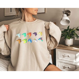 May include: A beige sweatshirt featuring a graphic print of 10 colourful dragon heads. The dragon heads are arranged in two rows of five. The dragon heads are all facing to the right.