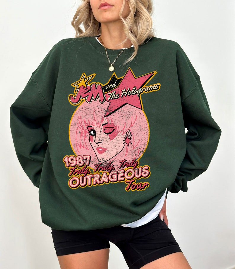 May include: Green sweatshirt with a vintage-style graphic of Jem and the Holograms. The graphic features a pink-haired Jem with a star on her ear, and the text "1987 Truly Outrageous Tour".