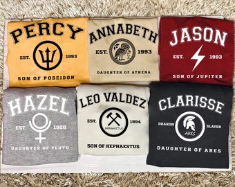 May include: Six different colored t-shirts with a graphic design and text. The shirts are all laid out on a beige carpet. The text on the shirts reads: Percy, Annabeth, Jason, Hazel, Leo Valdez, and Clarisse. The shirts are all different colors, including yellow, beige, red, gray, and black.