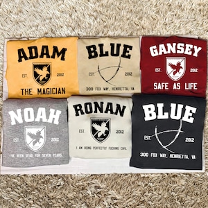 Puede incluir: Seis camisetas diferentes con varios diseños y textos. Las camisetas son todas negras, grises, blancas, beige, amarillas o rojas. El texto en las camisetas incluye "Adam", "Blue", "Gansey", "Noah", "Ronan" y "Safe as Life".