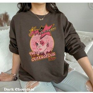 May include: A brown sweatshirt featuring a vintage-style graphic of Jem and the Holograms. The graphic depicts Jem's face against a pink background with the text "1987 Truly Truly Truly Outrageous Tour".