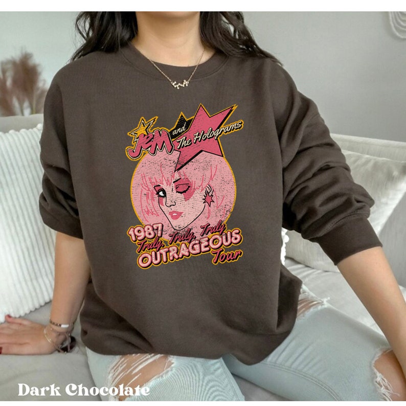 May include: A brown sweatshirt featuring a vintage-style graphic of Jem and the Holograms. The graphic depicts Jem's face against a pink background with the text "1987 Truly Truly Truly Outrageous Tour".