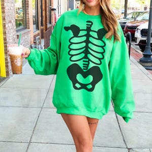 May include: A vibrant green sweatshirt with a black skeleton design. The sweatshirt has a crew neck and long sleeves. The image is taken outside on a sunny day.
