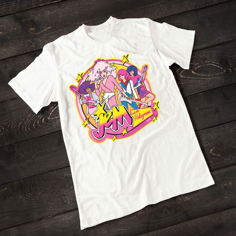 Może przedstawiać: Biała koszulka z r&oacute;żowym, ż&oacute;łtym i niebieskim nadrukiem przedstawiającym kresk&oacute;wkową grupę muzyczną Jem and the Holograms. Grafika przedstawia członk&oacute;w zespołu grających na instrumentach muzycznych oraz nazwę zespołu w stylizowanym foncie.