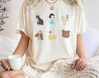 Könnte beinhalten: Cremefarbenes T-Shirt mit Cartoon-Illustrationen einer schwarzen Katze, eines Mädchens in einem blauen Kleid, eines Huhns, einer Eule, einer Ente und eines Bären. Das Shirt hat kurze Ärmel und eine lockere Passform.
