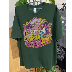 Może przedstawiać: Zielona koszulka z grafiką przedstawiającą zesp&oacute;ł kresk&oacute;wkowy Jem and the Holograms. Grafika przedstawia członk&oacute;w zespołu w ich charakterystycznych strojach i pozach, z nazwą zespołu w dużych, r&oacute;żowych literach.