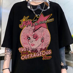 May include: Black t-shirt featuring a vintage-style graphic of Jem and the Holograms. The graphic depicts a pink-haired Jem with a star and the text "1987 Truly, Truly Outrageous Tour".