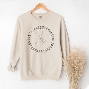 K&ouml;nnte beinhalten: Ein beiger Pullover mit einer schwarzen Grafik eines Kreises mit einem Kompass darin. Die Kompasspunkte sind mit den Buchstaben des Alphabets beschriftet.