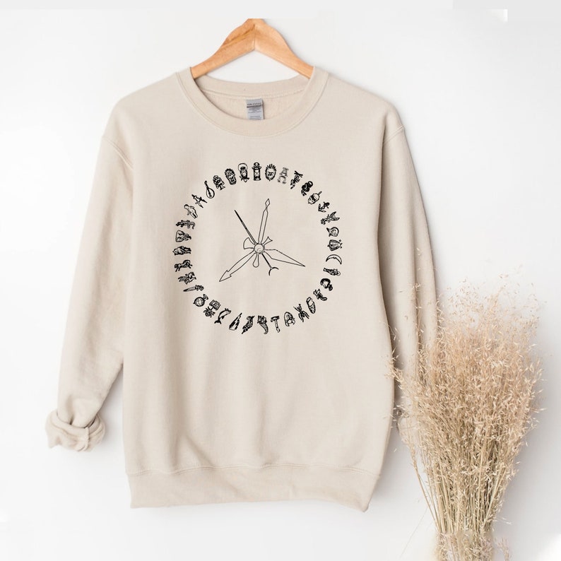 K&ouml;nnte beinhalten: Ein beiger Pullover mit einer schwarzen Grafik eines Kreises mit einem Kompass darin. Die Kompasspunkte sind mit den Buchstaben des Alphabets beschriftet.