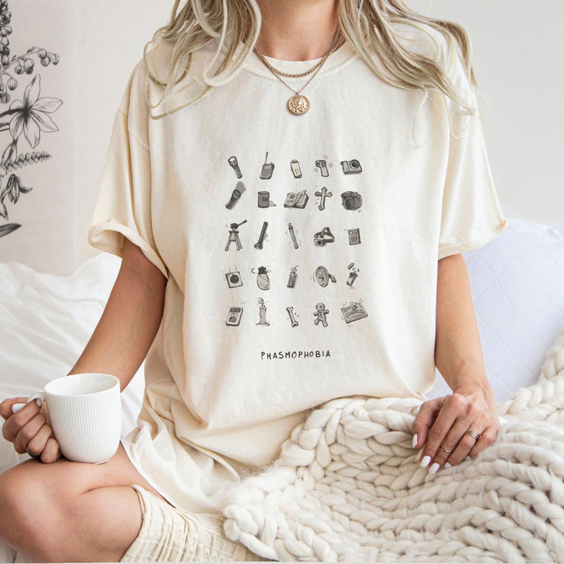 Puede incluir: Una camiseta blanca con un gr&aacute;fico en blanco y negro de varios art&iacute;culos utilizados en el videojuego "Phasmophobia". El texto "Phasmophobia" est&aacute; impreso debajo del gr&aacute;fico.