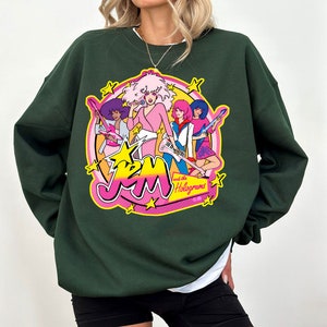 Może przedstawiać: Zielona bluza z nadrukiem graficznym przedstawiającym zesp&oacute;ł kresk&oacute;wkowy Jem and the Holograms. Nadruk przedstawia członk&oacute;w zespołu w ich charakterystycznych strojach i pozach, z nazwą zespołu i "and the Holograms" napisanymi r&oacute;żowymi i ż&oacute;łtymi literami.