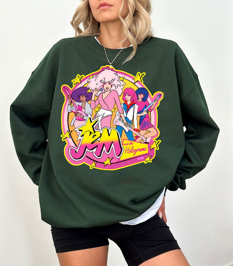 Może przedstawiać: Zielona bluza z nadrukiem graficznym przedstawiającym zesp&oacute;ł kresk&oacute;wkowy Jem and the Holograms. Nadruk przedstawia członk&oacute;w zespołu w ich charakterystycznych strojach i pozach, z nazwą zespołu i "and the Holograms" napisanymi r&oacute;żowymi i ż&oacute;łtymi literami.