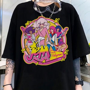 Może przedstawiać: Czarny T-shirt z nadrukiem graficznym zespołu kresk&oacute;wkowego Jem and the Holograms. Grafika przedstawia członk&oacute;w zespołu w ich charakterystycznych strojach i pozach, z ich imionami i nazwą zespołu w r&oacute;żowych i ż&oacute;łtych literach.