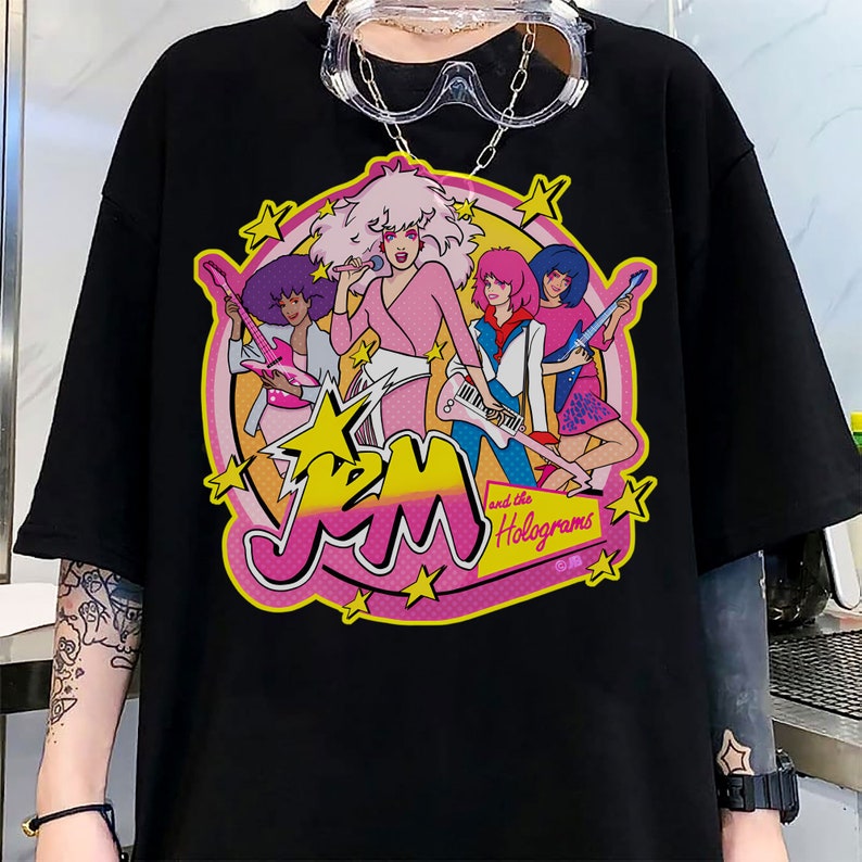 Może przedstawiać: Czarny T-shirt z nadrukiem graficznym zespołu kresk&oacute;wkowego Jem and the Holograms. Grafika przedstawia członk&oacute;w zespołu w ich charakterystycznych strojach i pozach, z ich imionami i nazwą zespołu w r&oacute;żowych i ż&oacute;łtych literach.