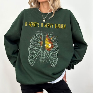 Op de afbeelding: Een donkergroen sweatshirt met de tekst "A heart's a heavy burden" en een afbeelding van een ribbenkast met een hart erin. Het hart is rood, geel en oranje.