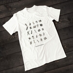 Puede incluir: Camiseta blanca con una ilustraci&oacute;n en blanco y negro de varios equipos de caza de fantasmas. El texto "PHASMOPHOBIA" est&aacute; impreso debajo de la ilustraci&oacute;n.