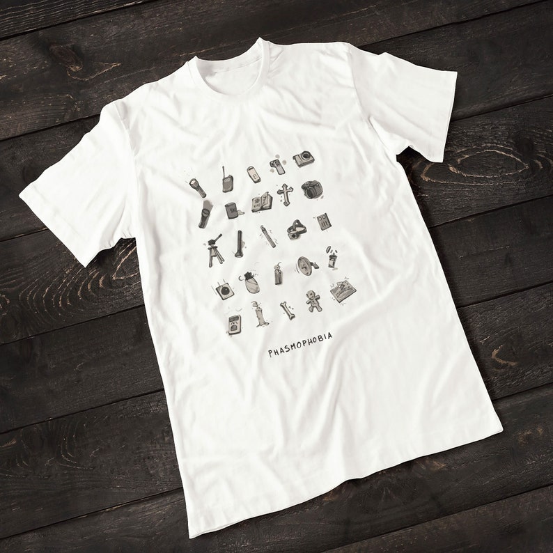 Puede incluir: Camiseta blanca con una ilustraci&oacute;n en blanco y negro de varios equipos de caza de fantasmas. El texto "PHASMOPHOBIA" est&aacute; impreso debajo de la ilustraci&oacute;n.