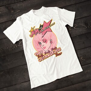 May include: White t-shirt with a vintage-style graphic of Jem and the Holograms. The graphic features a pink-haired Jem with a star and the text "1987 Truly Truly OUTRAGEOUS Tour".