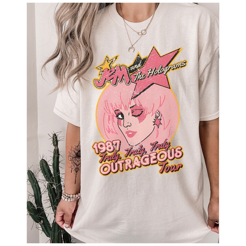 May include: White t-shirt featuring a pink and yellow graphic of Jem and the Holograms. The graphic includes the text "1987 Truly, Truly Outrageous Tour".