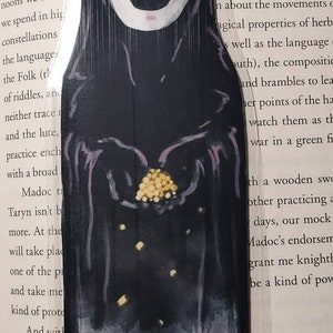 May include: A hand-painted bookmark featuring a black and white illustration of No Face from the movie Spirited Away. The bookmark is shaped like a person and has a white face with pink markings and black eyes. The character is holding a pile of gold coins in their hand.