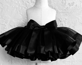 May include: A black tutu skirt with a large satin bow at the waist. The skirt is made of layers of tulle and has a satin ribbon trim along the bottom edge. The skirt is displayed on a mannequin.