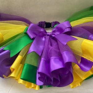 May include: A purple, green, and yellow tulle tutu with a purple satin bow. The tutu is layered with satin ribbon in each color.