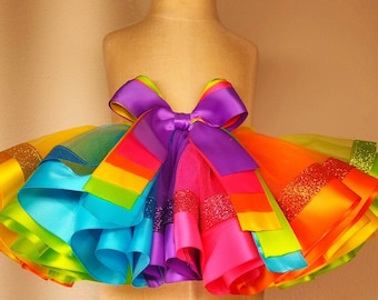 May include: A colorful rainbow tutu skirt with layers of tulle and satin ribbons. The skirt features a vibrant array of colors including yellow, green, blue, orange, pink, and purple. A large purple bow adorns the waistline, adding a decorative touch.