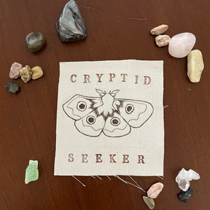 May include: A beige fabric patch with the words "CRYPTID SEEKER" embroidered in brown. A black and white illustration of a moth is centered on the patch.