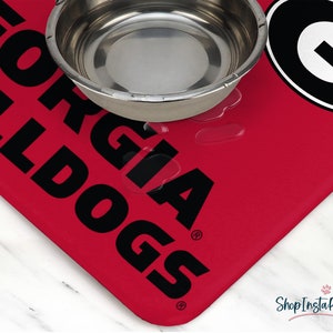 May include: A red pet food mat with the words "GEORGIA BULLDOGS" in large black letters. A stainless steel bowl sits on the mat, with water droplets visible. The mat has a rounded corner and the ShopInstaPets logo.