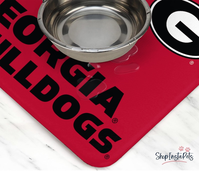 May include: A red pet food mat with the words "GEORGIA BULLDOGS" in large black letters. A stainless steel bowl sits on the mat, with water droplets visible. The mat has a rounded corner and the ShopInstaPets logo.