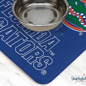 May include: Blue and white pet bowl mat with the University of Florida Gators logo and the words "Florida Gators" in white. The mat has a silver bowl on top of it.