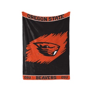 Puede incluir: Manta de forro polar negra con un logotipo naranja y negro de los Oregon State Beavers. El logotipo presenta una cabeza de castor con un diseño de pinceladas. El texto "OREGON STATE", "BEAVERS" y "OSU" está impreso en la manta.