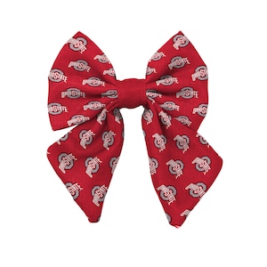 May include: A red fabric hair bow featuring a repeating pattern of the Ohio State University logo and text in white and grey. The bow has a structured, multi-layered design with a central knot and two long, pointed tails extending downwards.