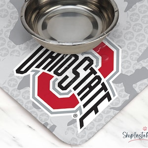 May include: A stainless steel pet bowl sits atop a gray and white mat featuring the Ohio State University logo in red, gray, and black. The mat has a repeating pattern of gray shapes and floral designs.