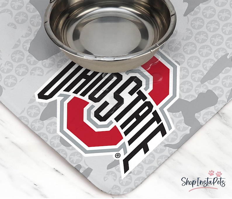 May include: A stainless steel pet bowl sits atop a gray and white mat featuring the Ohio State University logo in red, gray, and black. The mat has a repeating pattern of gray shapes and floral designs.