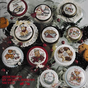May include: A collection of 10 round jar lids with cross-stitch designs featuring bears, hearts, and other cute motifs. The lids are decorated with lace and ribbons. The text on the lids includes "Happincas is Homemade", "A Gift From The Heart", "From My Kitchen To Yours", "My World Famous Chocolate Chip Cookies", "Made With Love", "You're The Best", "Thanks For Being A Friend", "Thank You", and "Thank You With All My Heart".