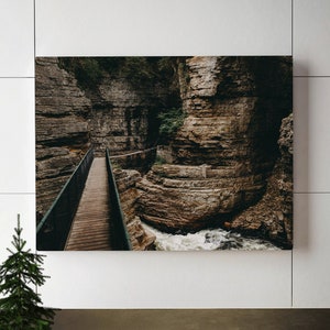 May include: A framed photograph of a wooden bridge over a rocky river in a canyon. The bridge is made of wood and metal and is surrounded by trees and rocks. The river is flowing rapidly and there is a waterfall in the background.