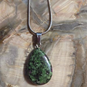 May include: A teardrop-shaped green and black gemstone pendant on a silver chain. The pendant has a mottled green and black pattern with hints of pink.