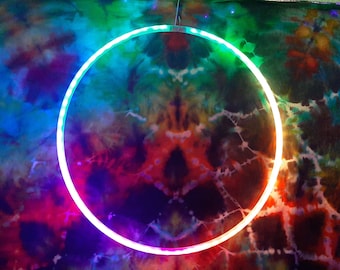 May include: A circular hoop featuring integrated LED lights that emit a vibrant gradient of colors, including blue, green, yellow, and red. The hoop is positioned against a tie-dye fabric background with a swirling pattern of dark and bright colors.