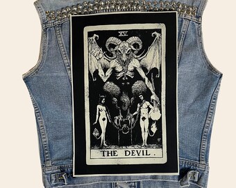 Puede incluir: Un chaleco vaquero con tachuelas plateadas y un parche negro con una carta del tarot que representa al Diablo. La carta es blanca sobre negra y muestra una figura con cuernos, alas y dos figuras encadenadas a ella. La carta dice "XV EL DIABLO."