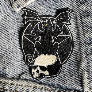 May include: Black embroidered patch of a cat with bat wings sitting on a skull with a pentagram in the background.