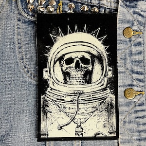 May include: Black and white patch of a skull wearing an astronaut helmet with a spiky design around the helmet. The patch is on a denim background.